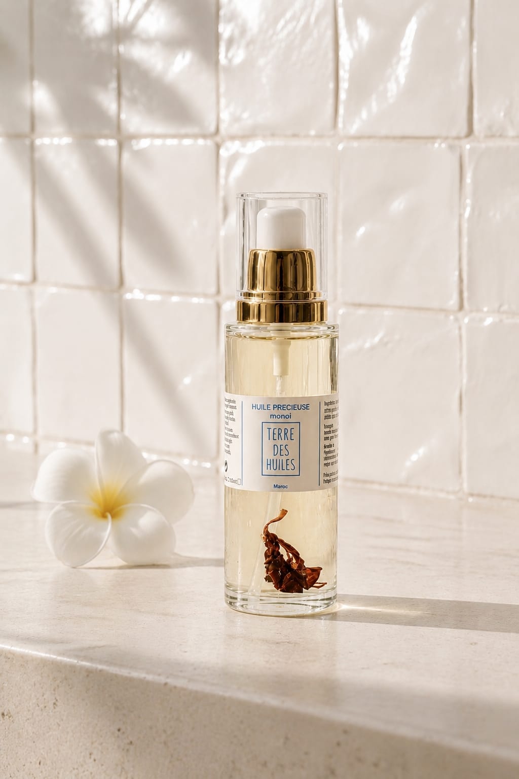 Flacon pompe en verre transparent de l'Huile Précieuse Monoï Terre Des Huiles, avec bague dorée et fleur de tiare à l'intérieur, posé sur un plan en pierre beige avec une fleur de frangipanier blanche, fond carrelage blanc texturé