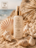 Spray solaire SPF50 Terre Des Huiles à l’huile d’argan sur plage avec coquillages et ambiance estivale premium.